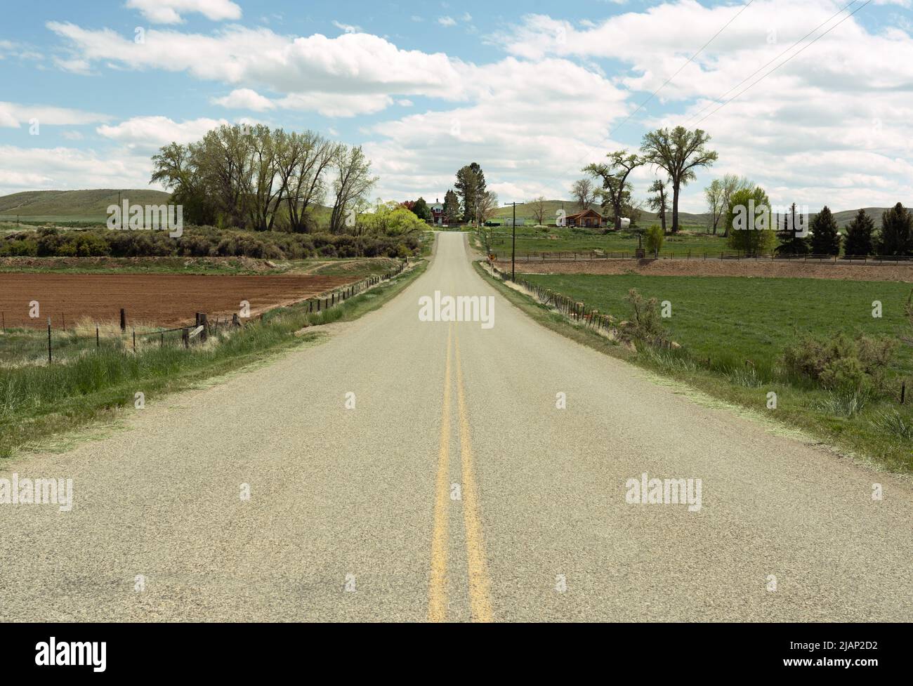 Guardando giù la linea gialla centrale di una strada a Shell, Wyoming si vede una fattoria in lontananza. Nuvole, cielo blu, alberi, pascoli sono inclusi. Foto Stock