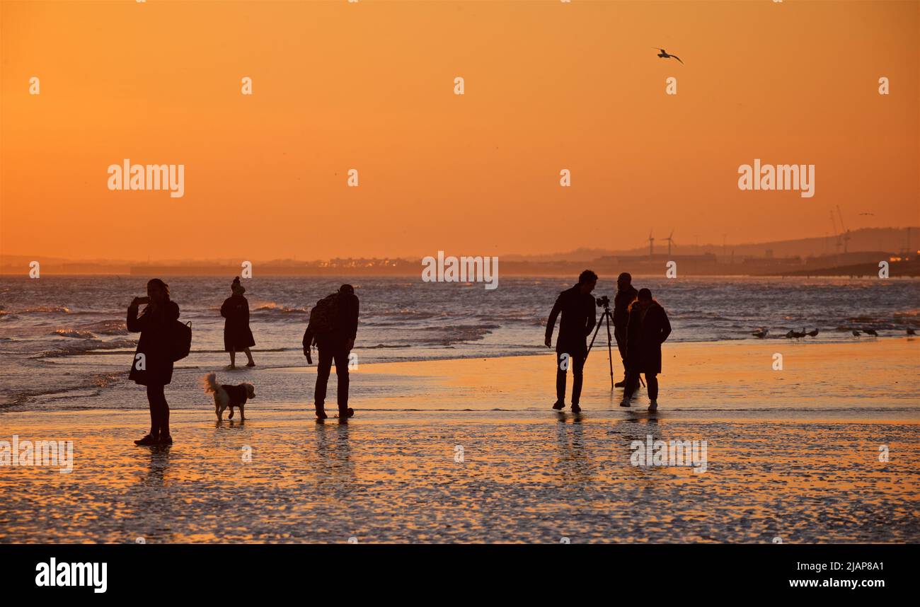 La fine della giornata ha plasmarsi le figure delle persone sulla spiaggia a bassa marea, Brighton & Hove, East Sussex, Inghilterra, Regno Unito. Fotografo con treppiede; cane. Guardando verso ovest verso Worthing Foto Stock