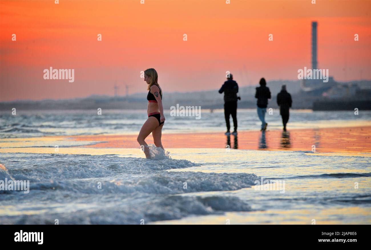 Forme di persone sulla spiaggia a bassa marea, Brighton & Hove, East Sussex, Inghilterra, Regno Unito. Donna in bikini che entra in mare . A marzo, le temperature dell'acqua sono generalmente inferiori a 10 gradi celsius. Foto Stock
