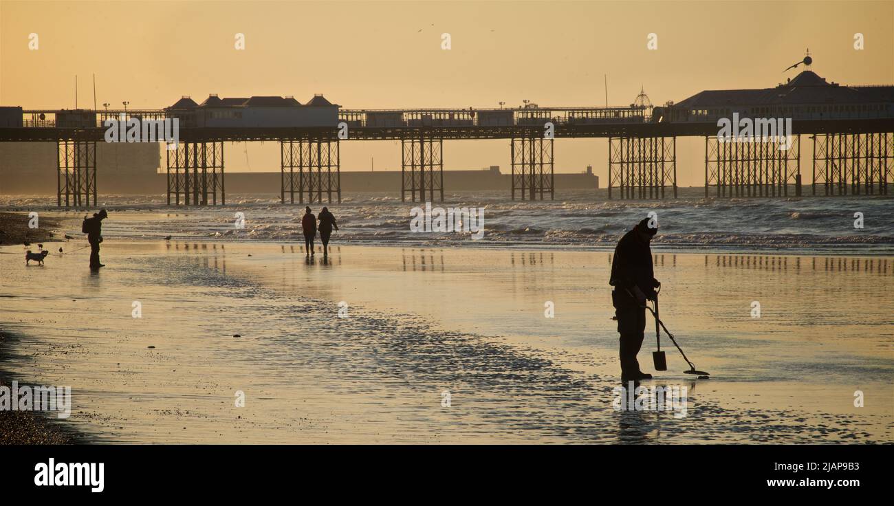 Metal detectorist prospezione sulla sabbia a basso tempo. Spiaggia all'alba, Brighton & Hove, Sussex, Inghilterra, Regno Unito. PPeople camminando sulla sabbia, alcuni con un cane; Palace Pier oltre. Foto Stock