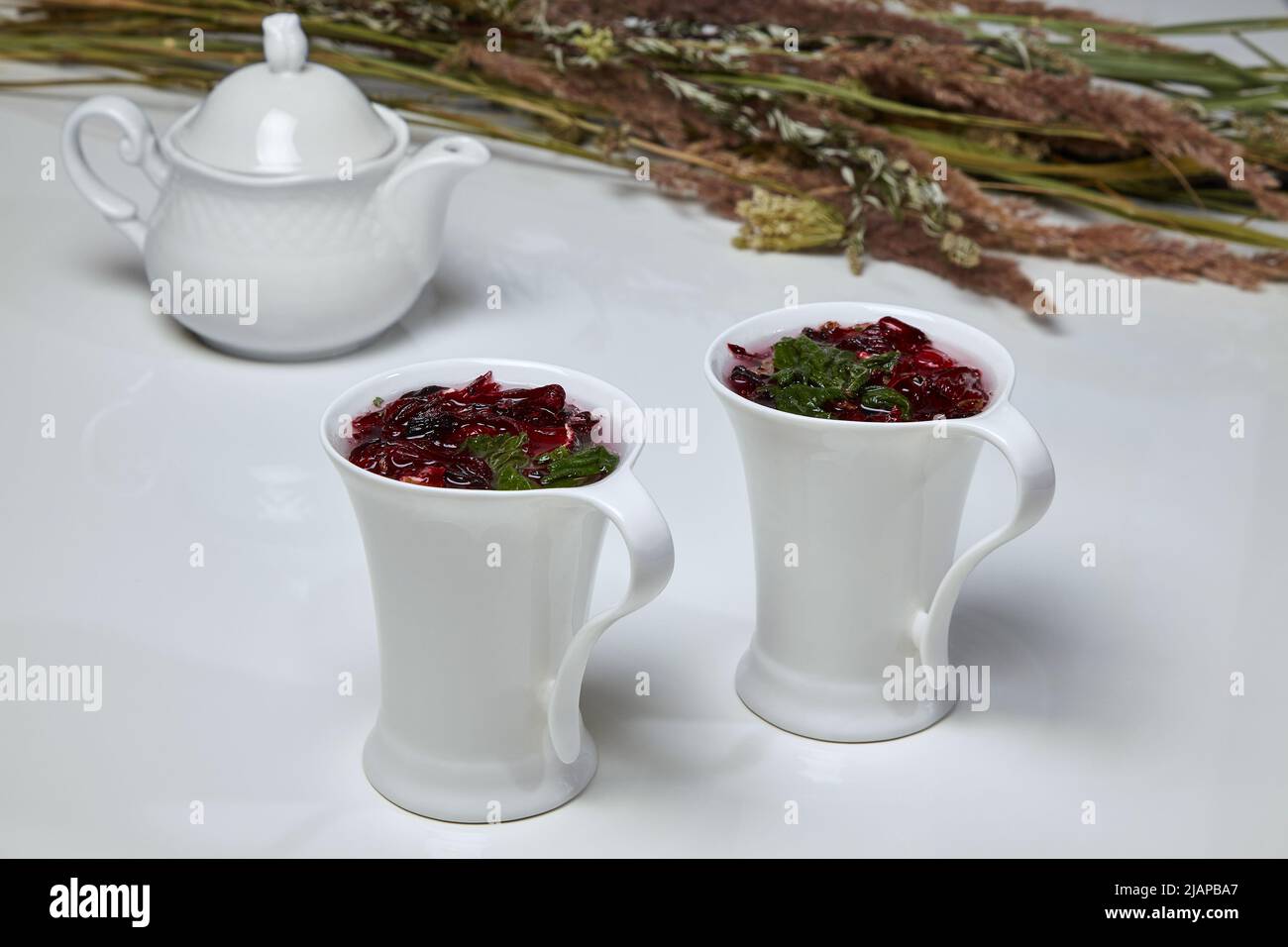 Il tè alle erbe di Carcade è composto da fiori di rosa e menta sudanese in eleganti tazze bianche su un tavolo bianco sullo sfondo di una teiera e un bou Foto Stock