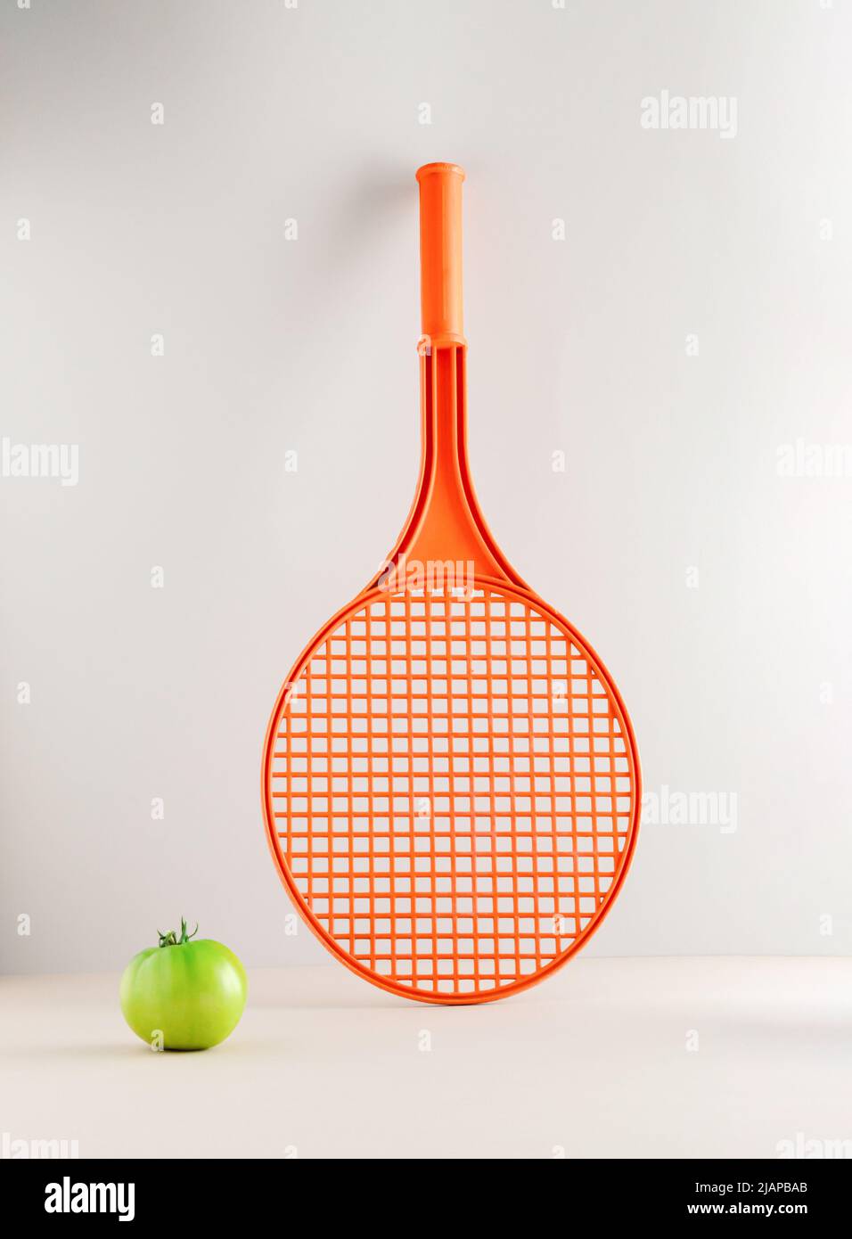 Una racchetta da tennis e un pomodoro verde non maturo su sfondo grigio. Concetto sportivo minimo. Foto Stock