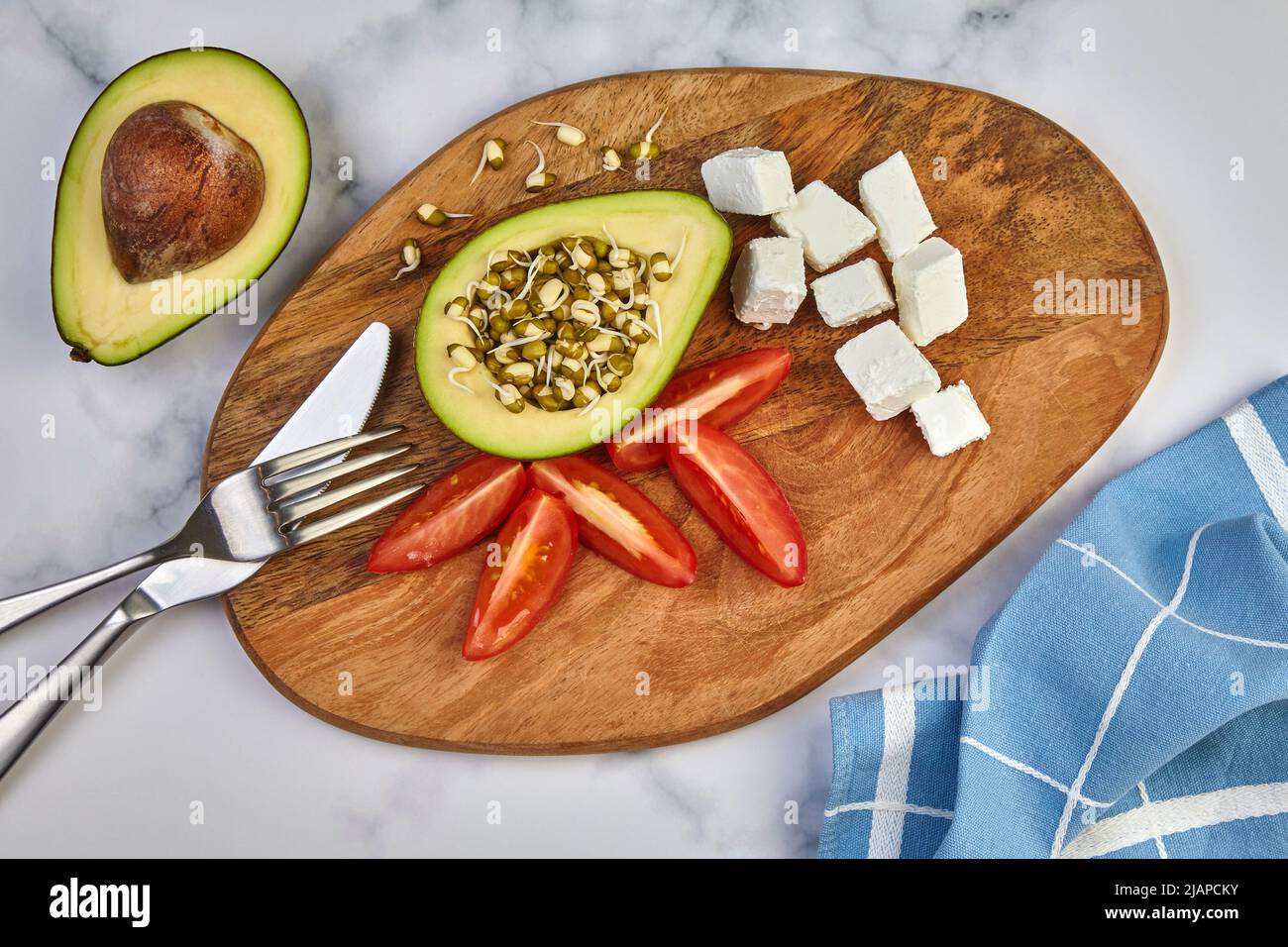 Avocado germogli mungo fagiolo pomodori cagliati posate di formaggio e un asciugamano da cucina su un asse di legno su un tavolo leggero. Vista dall'alto Foto Stock