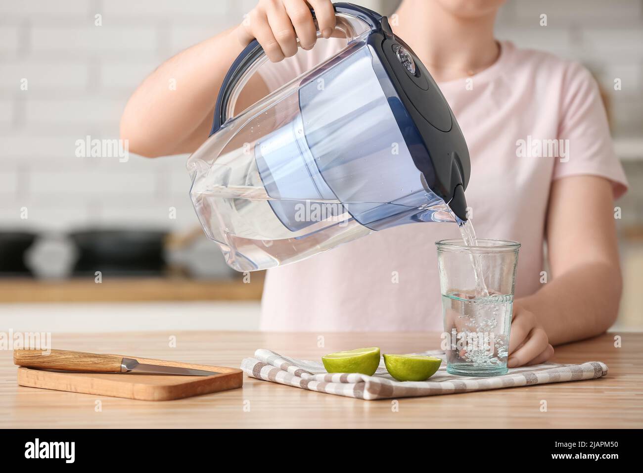 Donna che versa acqua purificata in vetro dal contenitore del filtro al tavolo in cucina Foto Stock