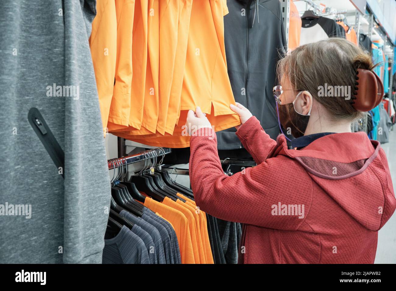 Donna caucasica in maschera contro il virus sceglie una camicia arancione per acquistare in un negozio Foto Stock