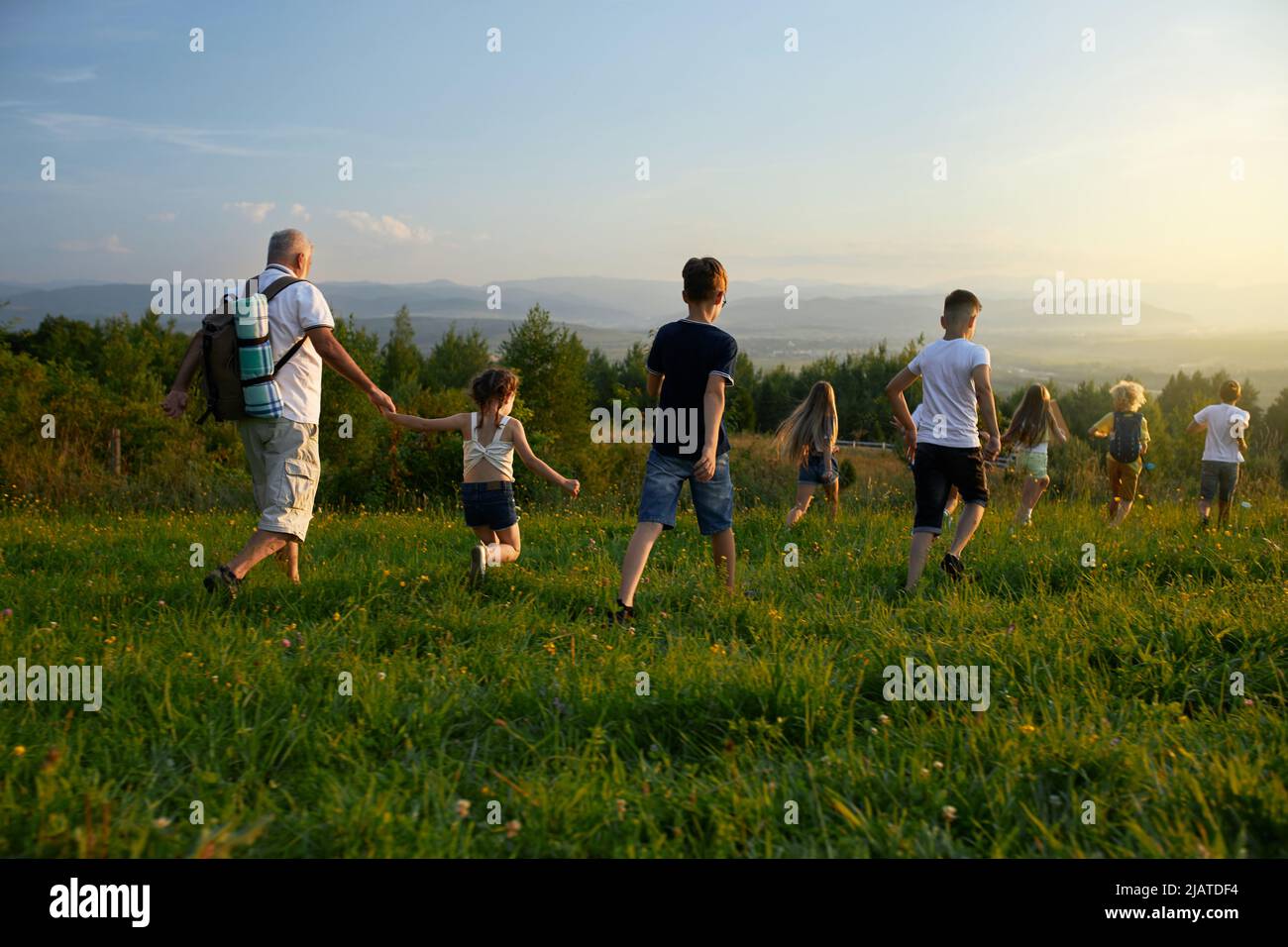 Vista posteriore dei bambini con il vecchio uomo che si guida in avanti in collina. Maschio con capelli grigi che indossano zaino insieme ai bambini che camminano, viaggiano, corrono avanti. Concep di riposo attivo. Foto Stock