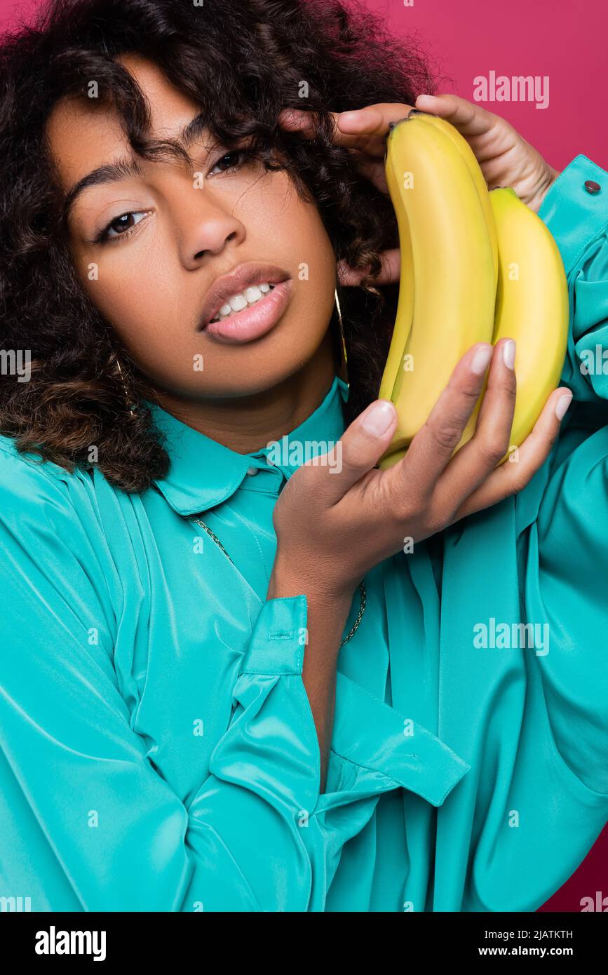 bella donna afroamericana in blusa blu che tiene banane e guarda la macchina fotografica isolato su rosa Foto Stock