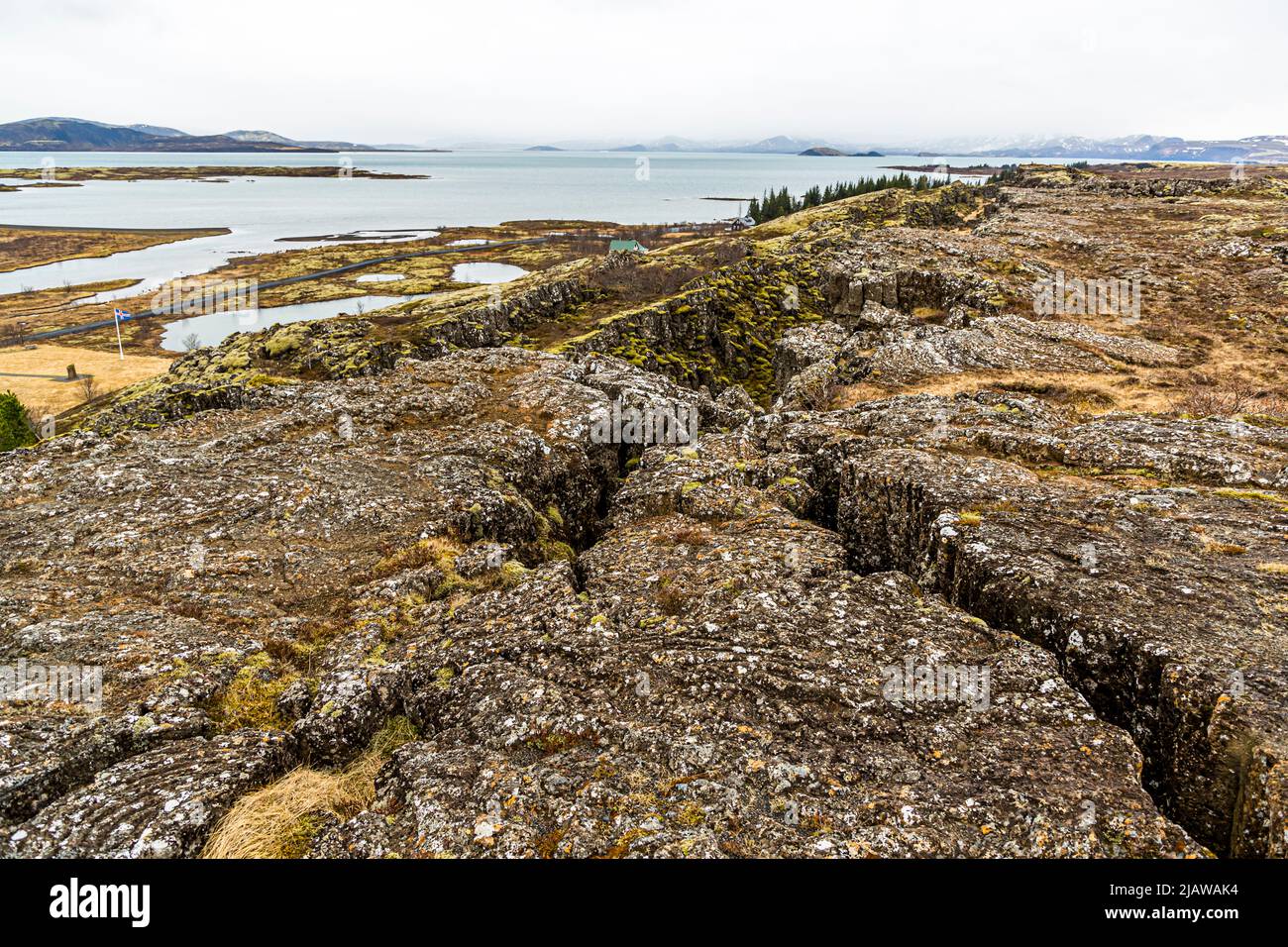 La pianura di lava strappata di Þingvellir, in Islanda, si divide in profonde fessure e crepacci che mostrano la frattura della crosta terrestre lungo il confine tra le placche nordamericana ed eurasiatica. Dietro si trova il lago Þingvallavatn, il più grande lago naturale del paese, incorniciato da montagne innevate. La bandiera nazionale sventola sopra il sito di assemblea al bordo sinistro dell'immagine. Intorno al 930 gli islandesi fondarono qui la loro assemblea generale Alþing, che si riunì fino al 1798, e l'area è stata patrimonio mondiale dell'UNESCO dal 2004. Foto Stock