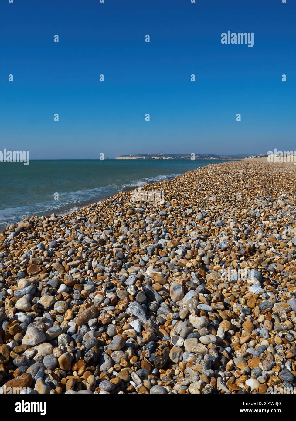 Una scena marina illuminata dal sole a Seaford, guardando attraverso l'ampia spiaggia di ghiaia e le onde del canale Inglese fino al promontorio attraverso la baia. Foto Stock