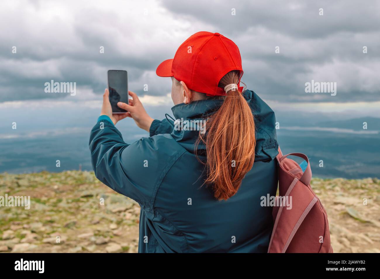 Giovane caucasica 30s donna con zaino in berretto rosso scattando una foto sulla cima delle montagne. Vista posteriore Foto Stock