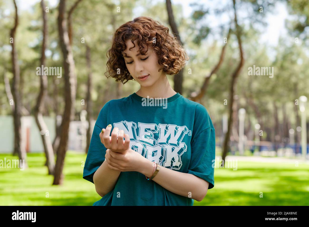 Giovane donna rossa che indossa una t-shirt verde in piedi sul parco cittadino, all'aperto tenendo il suo polso doloroso. Dolore sulle mani e sulle dita, artrite dentro Foto Stock