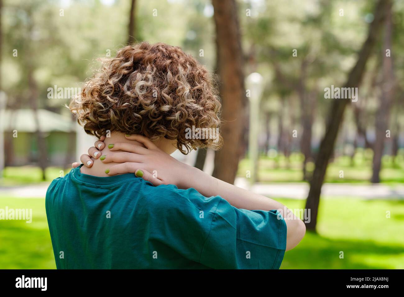 Vista posteriore della giovane donna rossa che indossa il tee verde in piedi sul parco della città, all'aperto sensazione di dolore alle spalle articolare dolore alla schiena, concetto di fibromialgia, cl Foto Stock