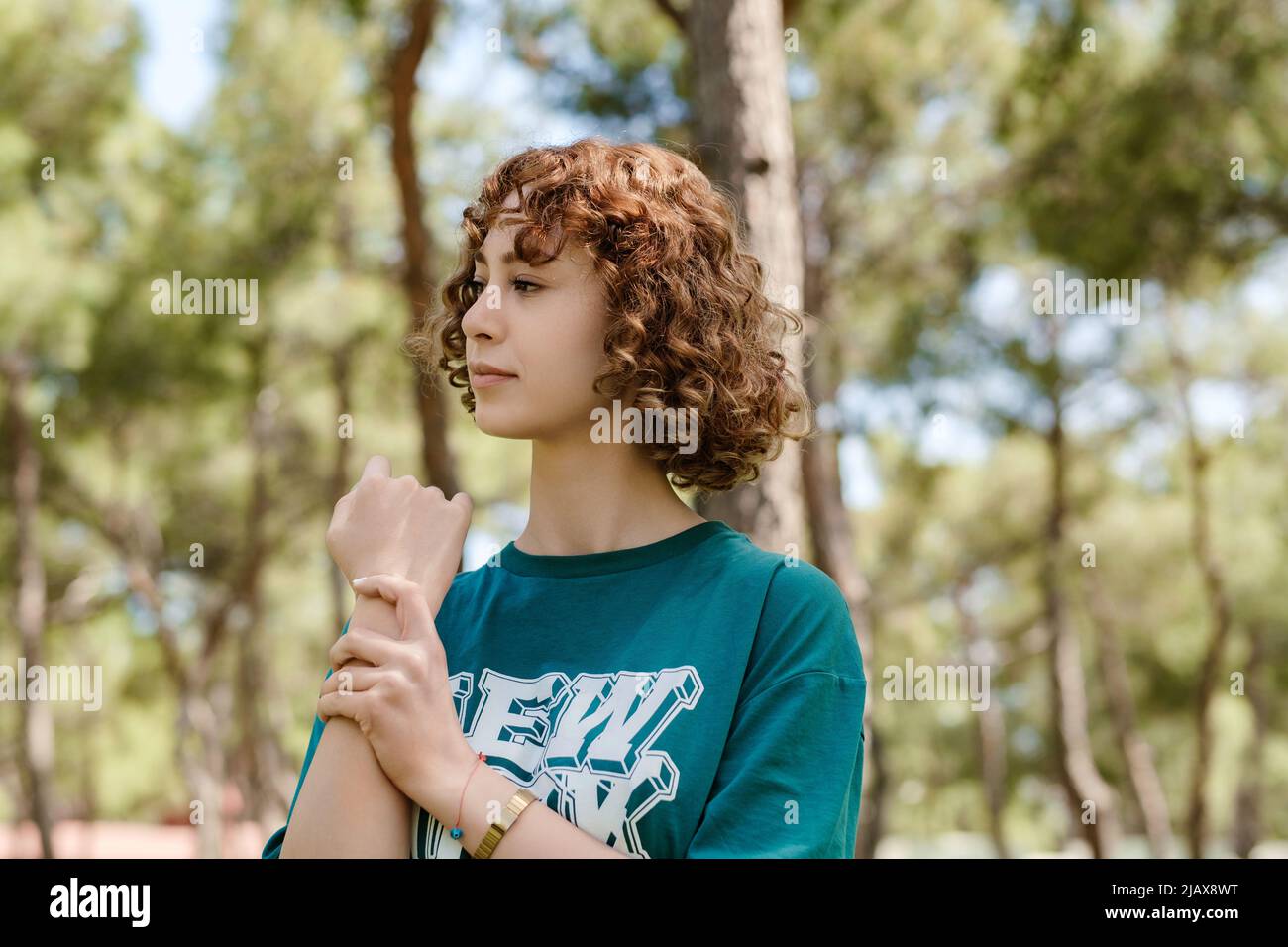 Giovane donna che indossa una t-shirt verde casual in piedi sul parco della città, guardando fuori mentre tiene il suo polso doloroso. Dolore alle mani e alla pinna Foto Stock