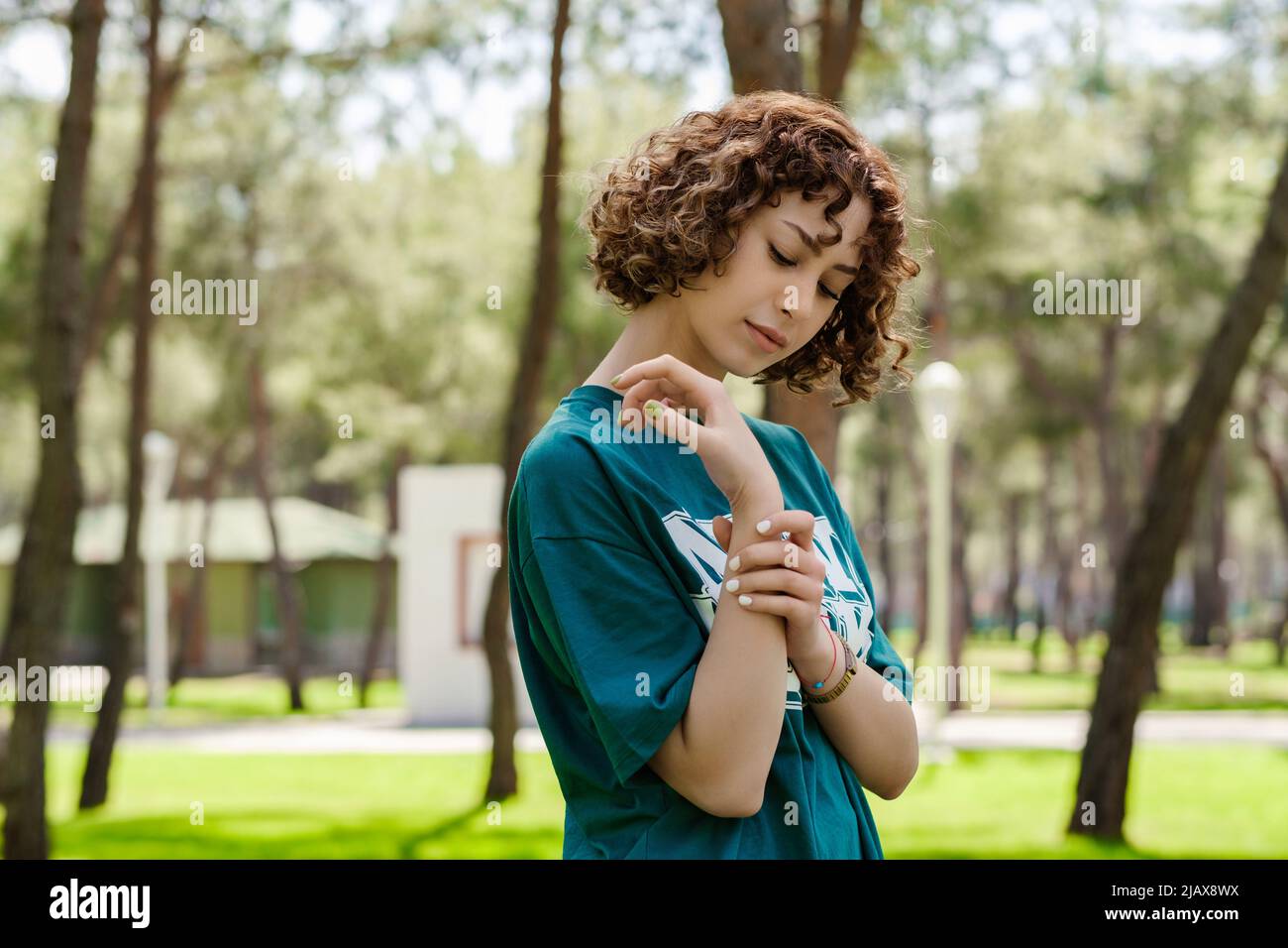 Vista laterale della giovane donna triste con t-shirt verde in piedi sul parco cittadino, all'aperto tenendo il suo polso doloroso. Dolore alle mani e alle dita, arte Foto Stock