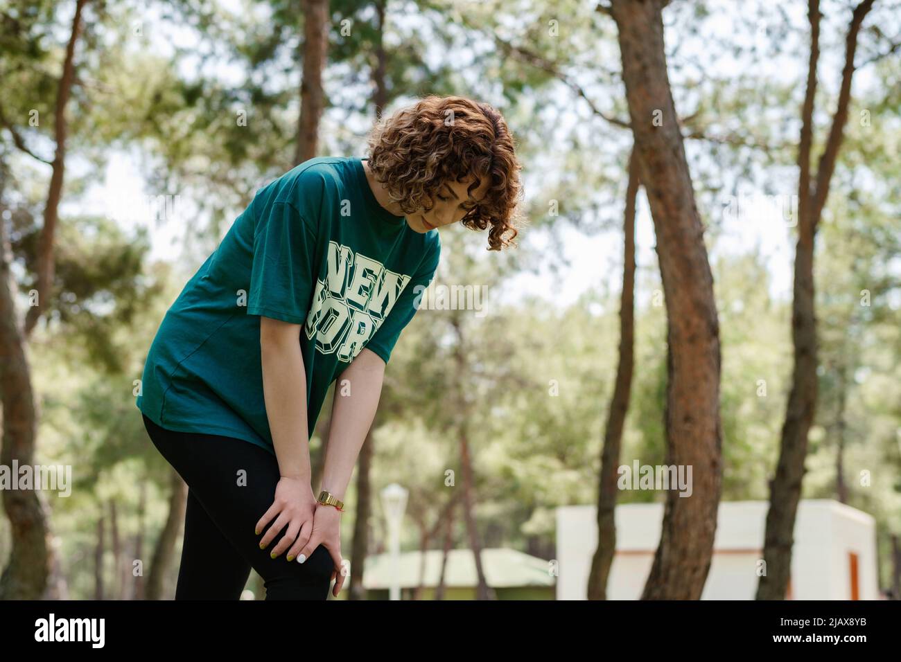 Vista laterale di una giovane donna rossa che indossa abiti sportivi in piedi sul parco cittadino, all'aperto toccando il ginocchio ferito con le mani. Gamba ferita, healt Foto Stock