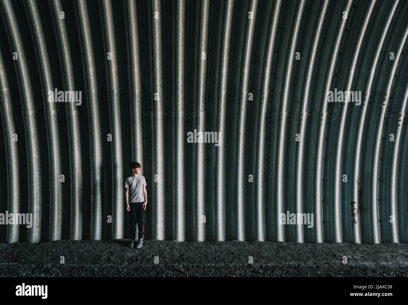 Ragazzo in piedi da solo in grande tunnel di metallo rigato guardando verso l'esterno. Foto Stock
