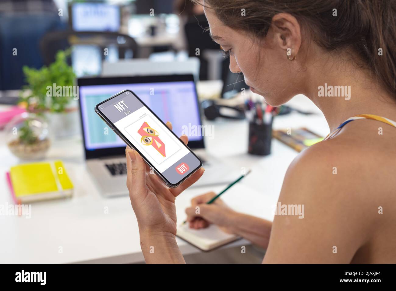 Giovane donna d'affari caucasica che scrive sul diario mentre usa lo smart phone alla scrivania in ufficio Foto Stock