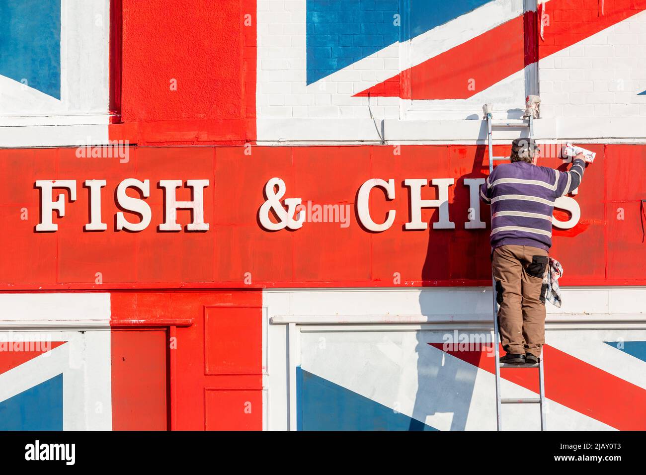 Cradley Heath, West Midlands, Regno Unito. 1st giugno 2022. Warren Rudge, il proprietario del chip shop, mette i tocchi finali sulla Union Jackflag che lui e suo figlio James hanno dipinto sul loro negozio di pesci e chip a Cradley Heath, West Midlands. La bandiera dipinta copre tutta la facciata di Ivan Fish and Chips, comprese le finestre, e ha preso tutto il giorno per completare. 'Ivan's Fish and Chips serve pesce e patatine da 65 anni, ma non tanto quanto sua Maestà. Ma ho sentito dire che la regina è parziale a fish and chips." dice Warren. Credit: Peter Lopeman/Alamy Live News Foto Stock