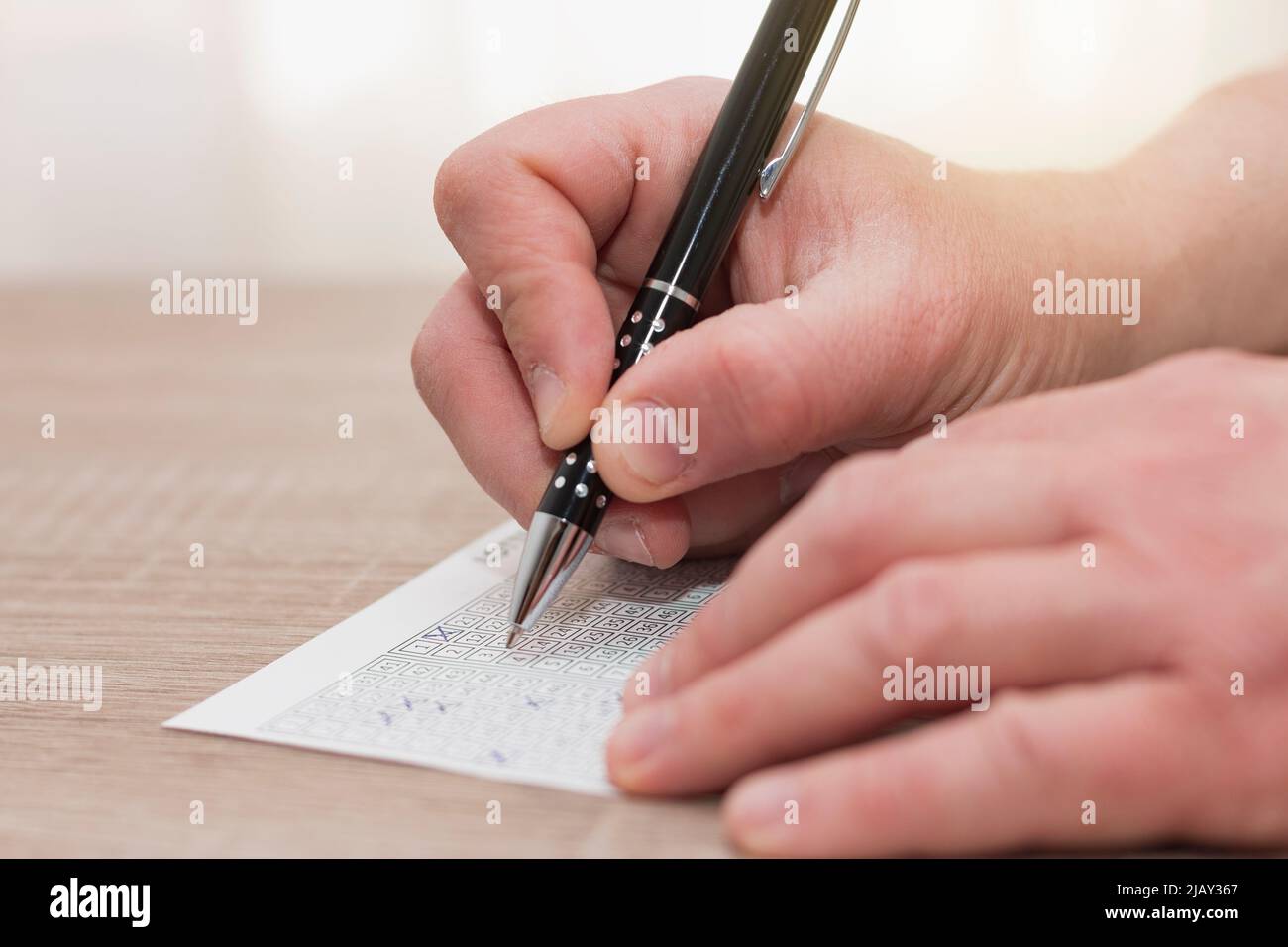La mano di un uomo caucasico irriconoscibile che tiene una penna nera mentre i numeri marcano su un biglietto della lotteria su un tavolo di legno. Foto Stock