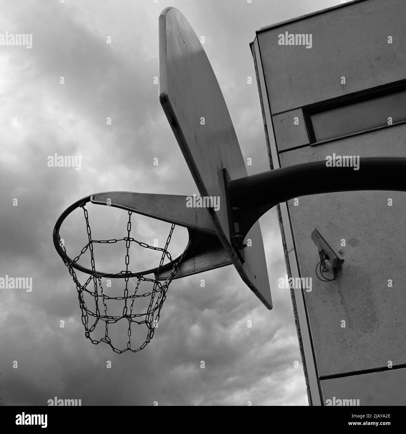 un cerchio da basket antivandalo con catene di ferro contro un cielo oscuro e nuvoloso, accanto ad un edificio in cemento con una telecamera di sorveglianza sul muro Foto Stock