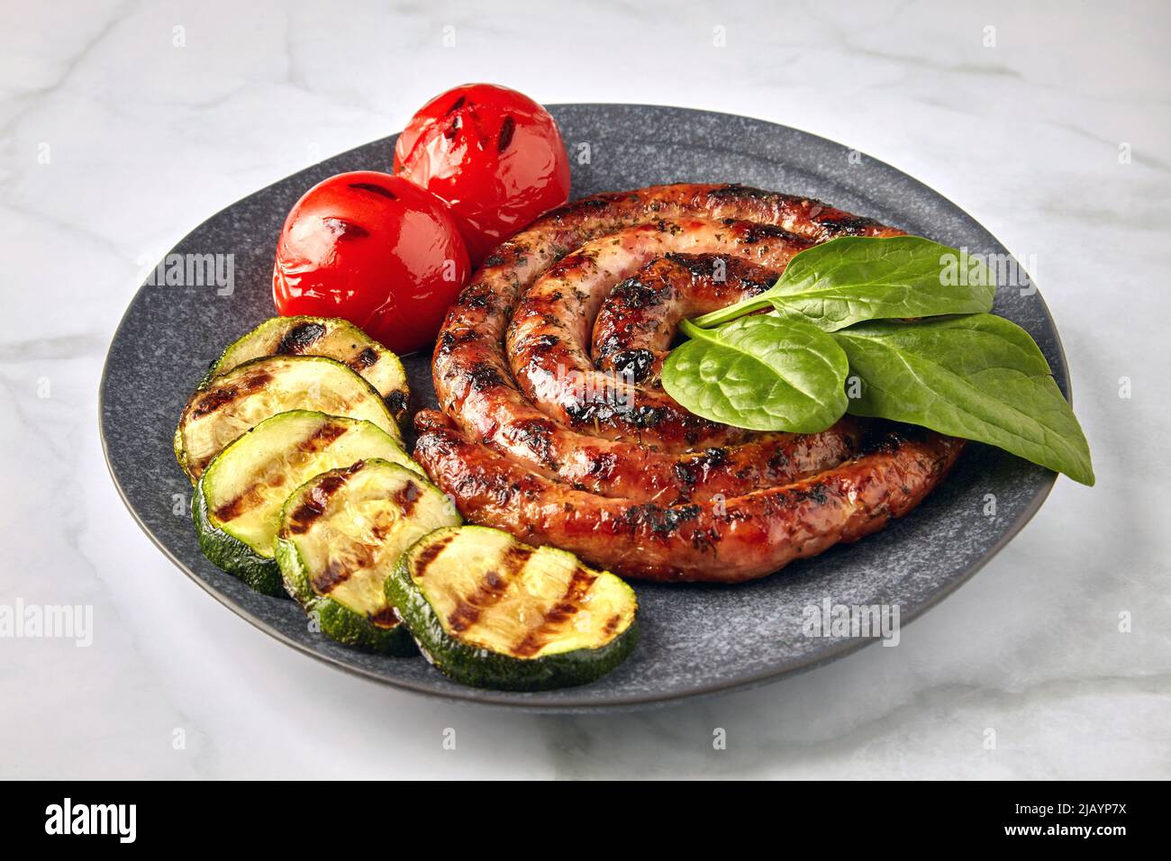 Salsiccia a spirale grigliata con zucchine, pomodori e basilico su un piatto scuro su un tavolo chiaro Foto Stock