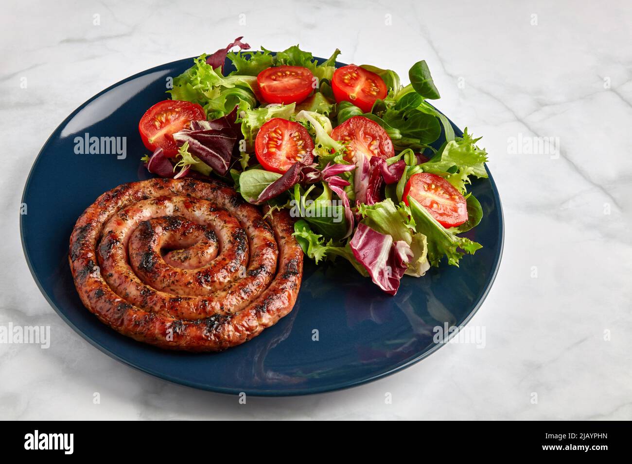 Salsiccia alla griglia con pomodori e erbe tritati su un piatto blu su un tavolo leggero Foto Stock