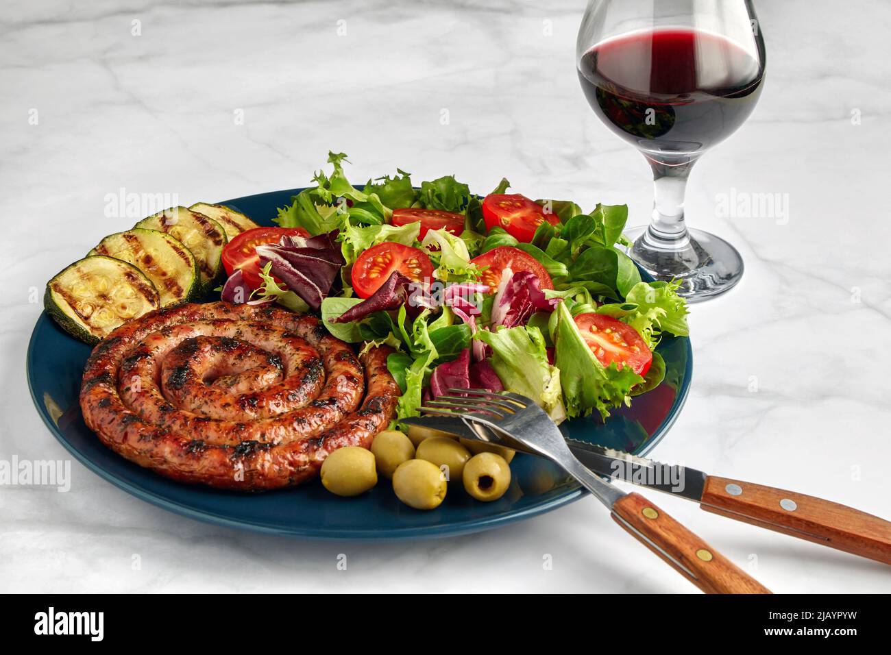 Salsiccia alla griglia con pomodori e erbe tritati su un piatto blu con un bicchiere di vino rosso su un tavolo leggero Foto Stock