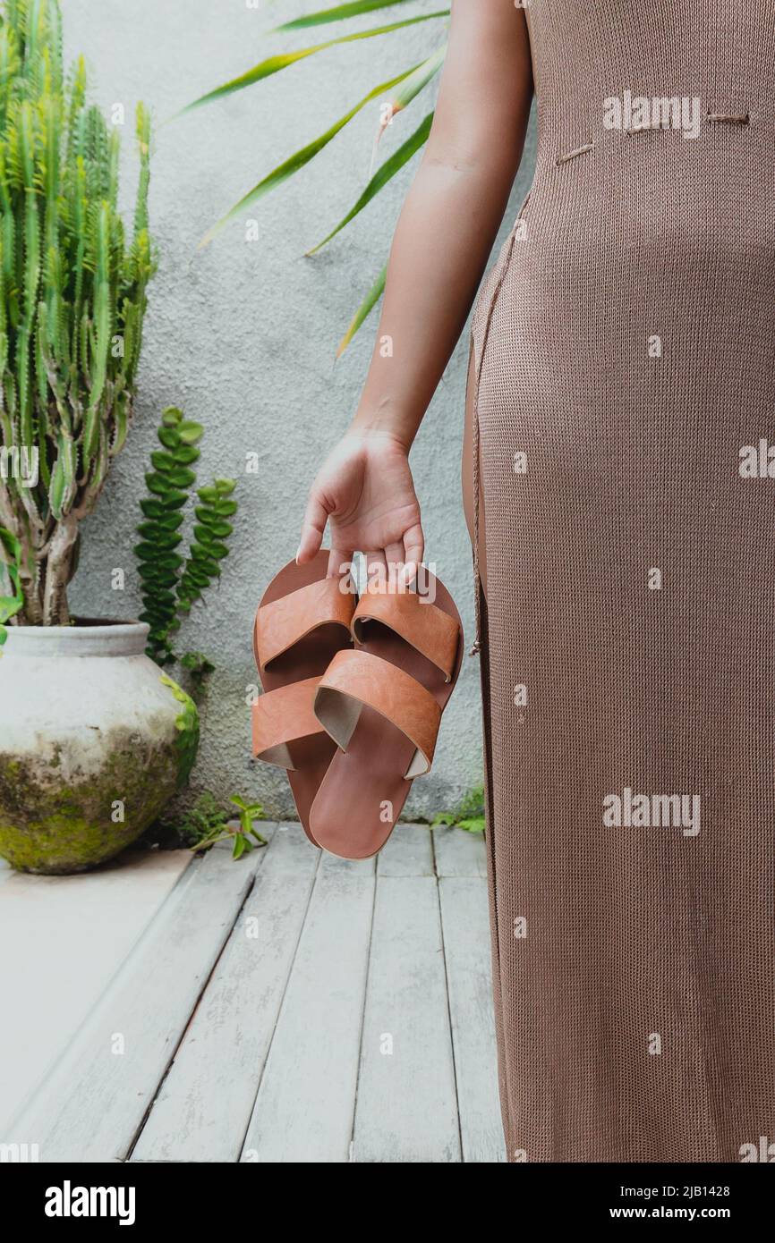 donna che tiene un paio di sandali marroni nel patio in un lungo vestito marrone con piante verdi sullo sfondo Foto Stock