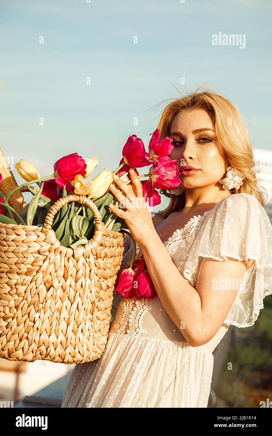 Ritratto di giovane donna con capelli lunghi e leali vestito bianco, orecchini floreali, in piedi vicino sacchetto di paglia con bouquet. Foto Stock