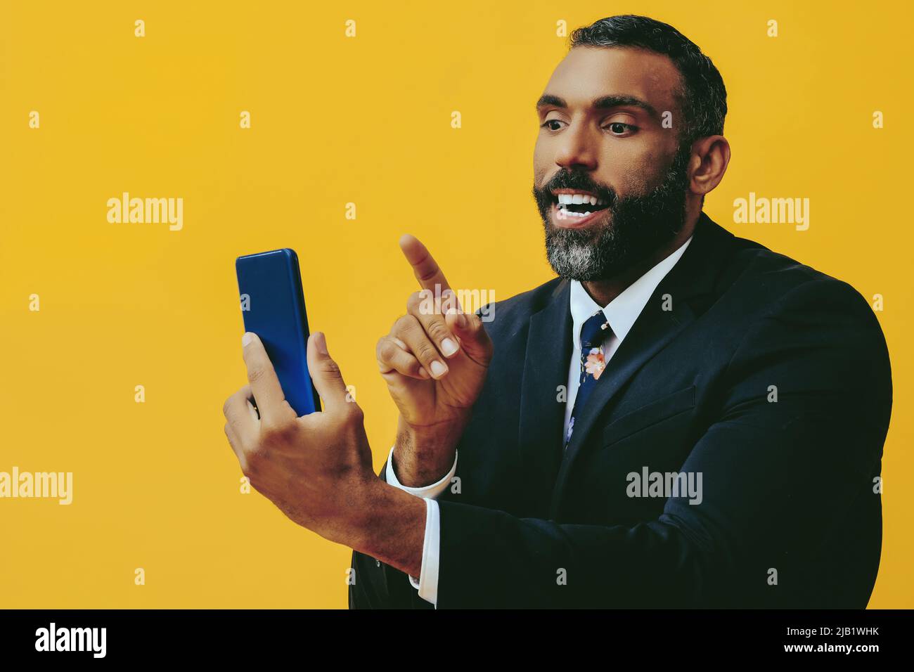 ritratto di sorridente positivo bel bearded uomo in vestito e legare con smartphone video chiamata che punta su sfondo giallo studio Foto Stock