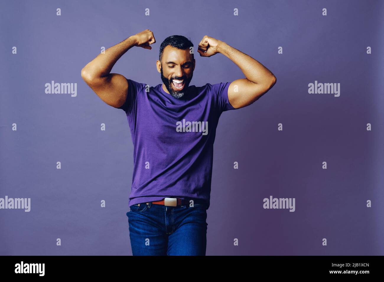 ritratto di un uomo vincitore di successo con una barba e una t-shirt viola che festeggia con il braccio su uno studio di sfondo viola Foto Stock