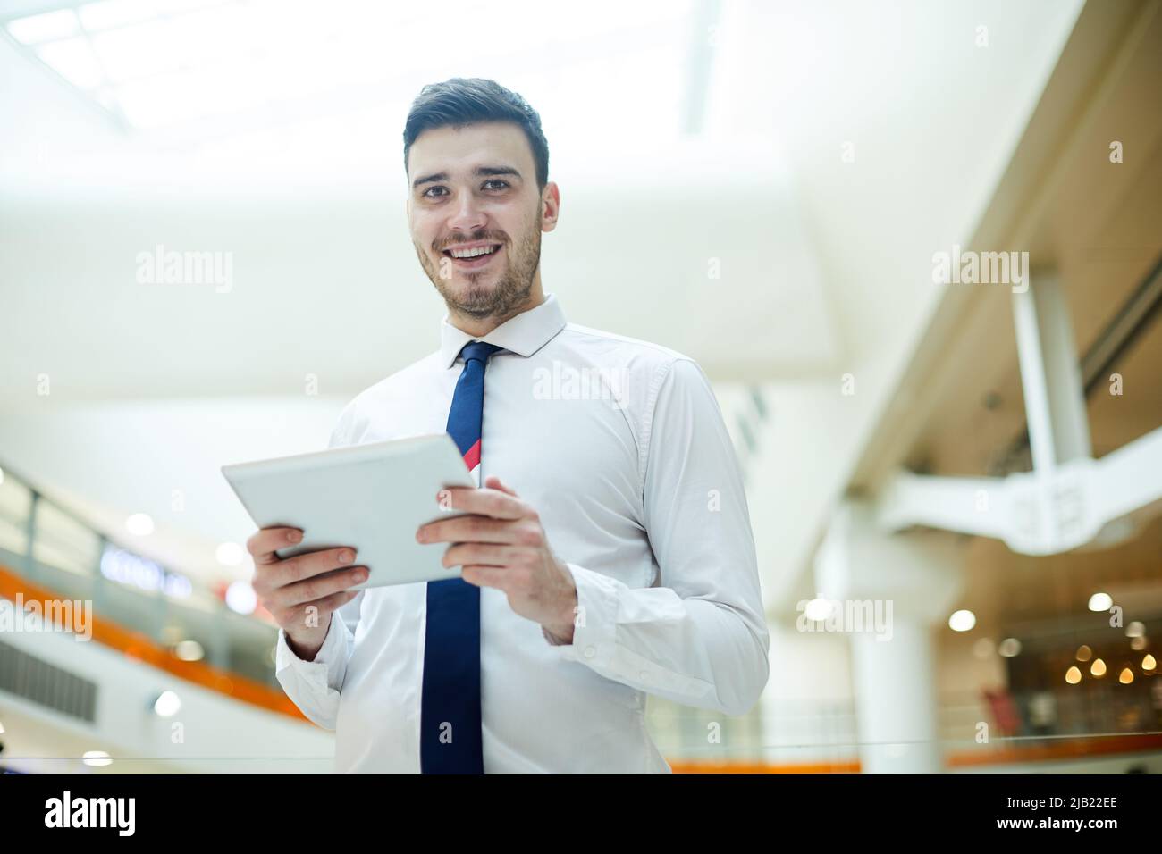 Sorridente e ottimista, giovane manager maschile con stubbie in piedi nella hall e utilizzando il tablet digitale durante l'analisi dei dati online Foto Stock