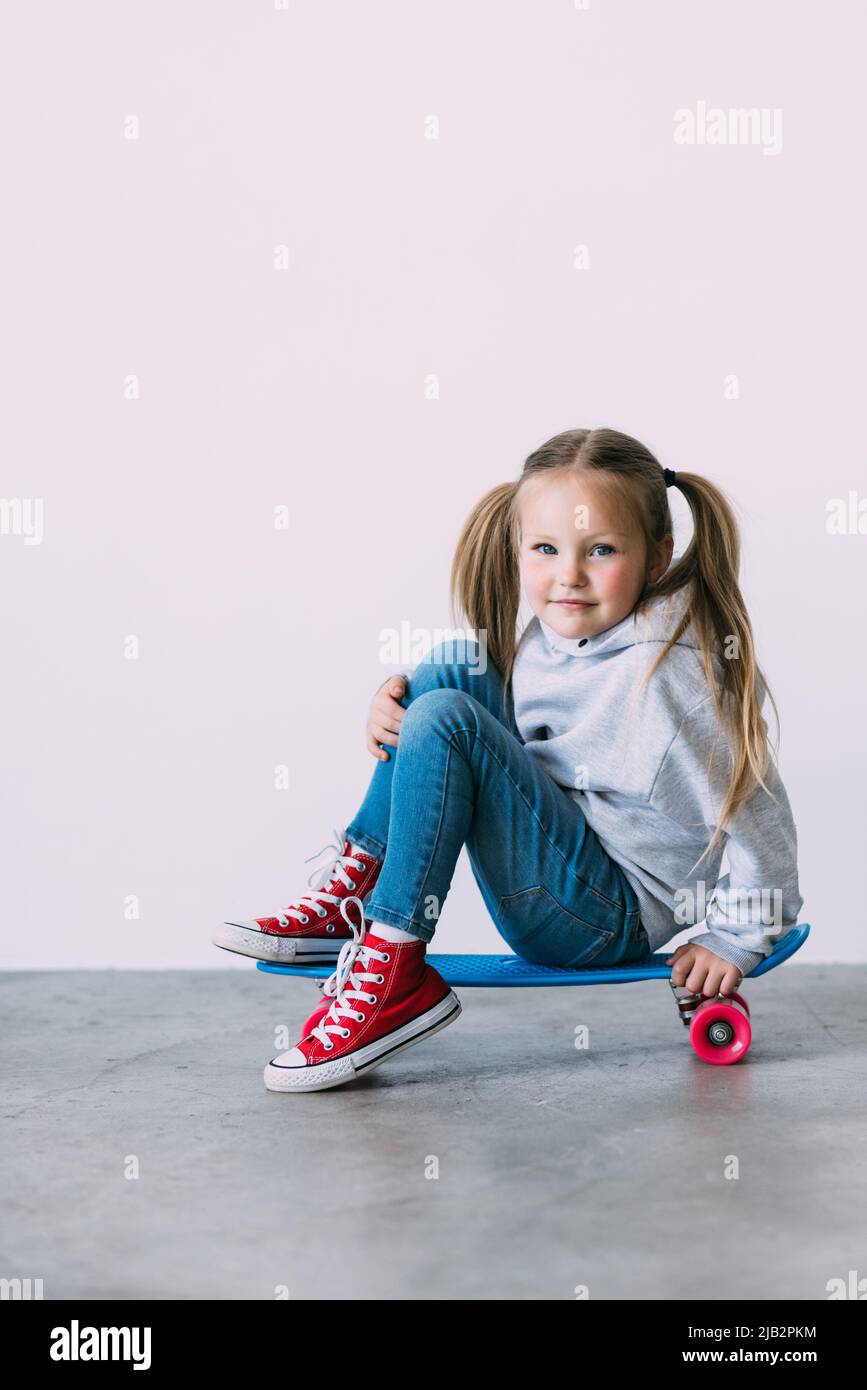 Sorridendo bambina in un abito estivo, seduto su uno skateboard, e si gonna una testa una mano, guardando la macchina fotografica con soddisfazione e happine Foto Stock