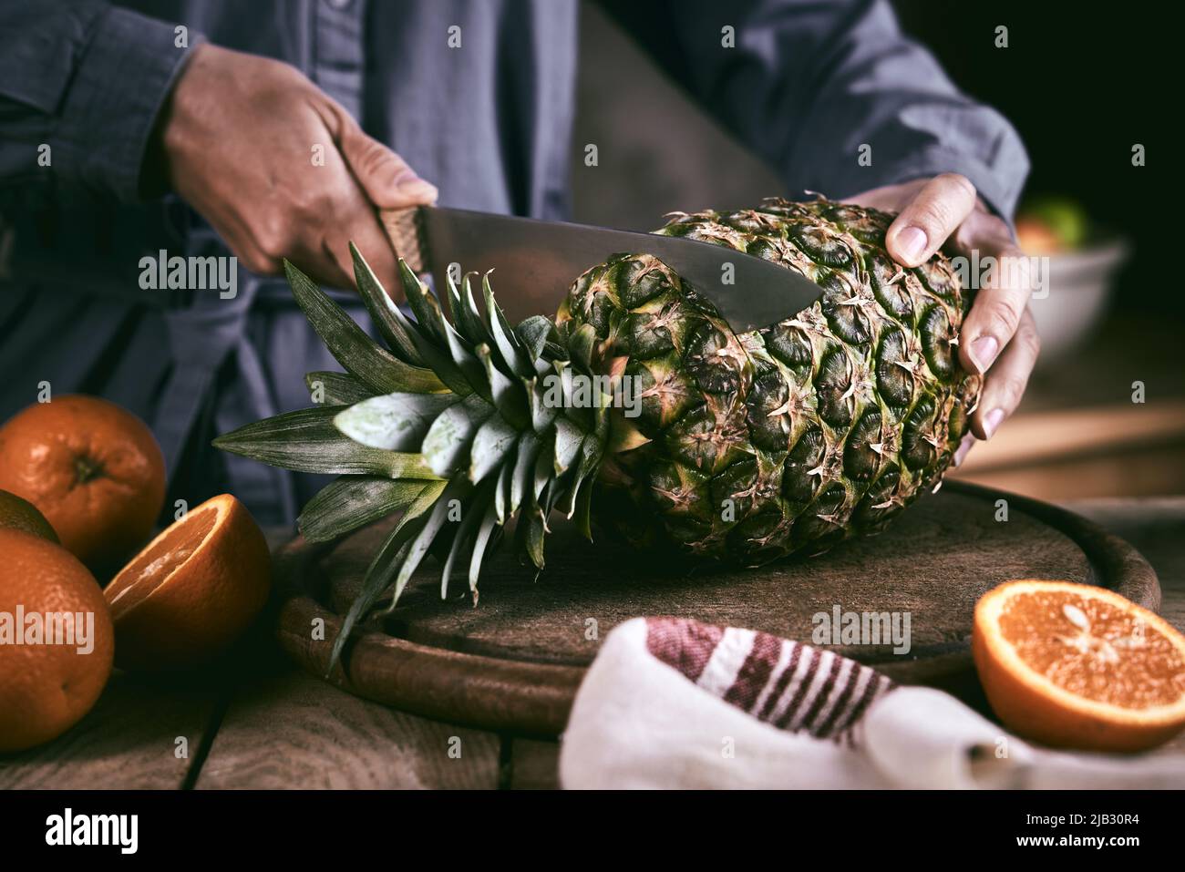 Donna irriconoscibile in camicia blu con coltello affilato per rimuovere le foglie di ananas fresco vicino arance mature sul tavolo in cucina a casa Foto Stock
