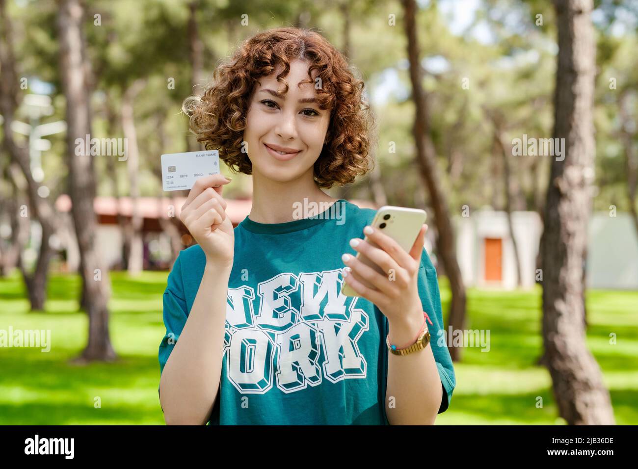 Giovane donna rossa che indossa una t-shirt verde in piedi sul parco cittadino, all'aperto mostrando carta di credito o debito plastica mentre tiene il telefono cellulare. Negozio online Foto Stock