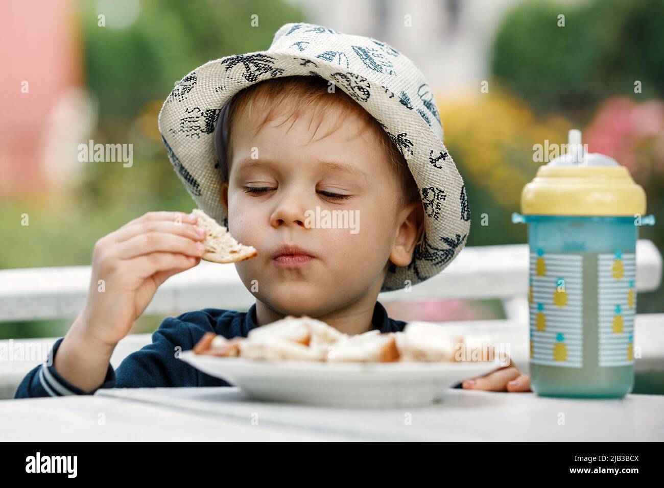 Un bambino sano mangia il burro sul pane nel giardino della nonna durante le vacanze estive. Una tazza di plastica con succo di ananas sul tavolo. Il conc Foto Stock