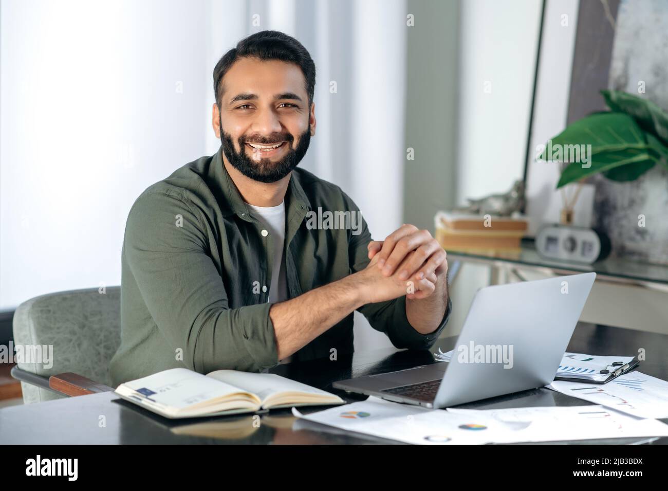 Ritratto di un bel positivo di successo arabo o indiano uomo vestito in abiti casual elegante, esperto finanziario, commerciante di stock, seduto alla sua scrivania, guardando la macchina fotografica, sorridente amichevole Foto Stock