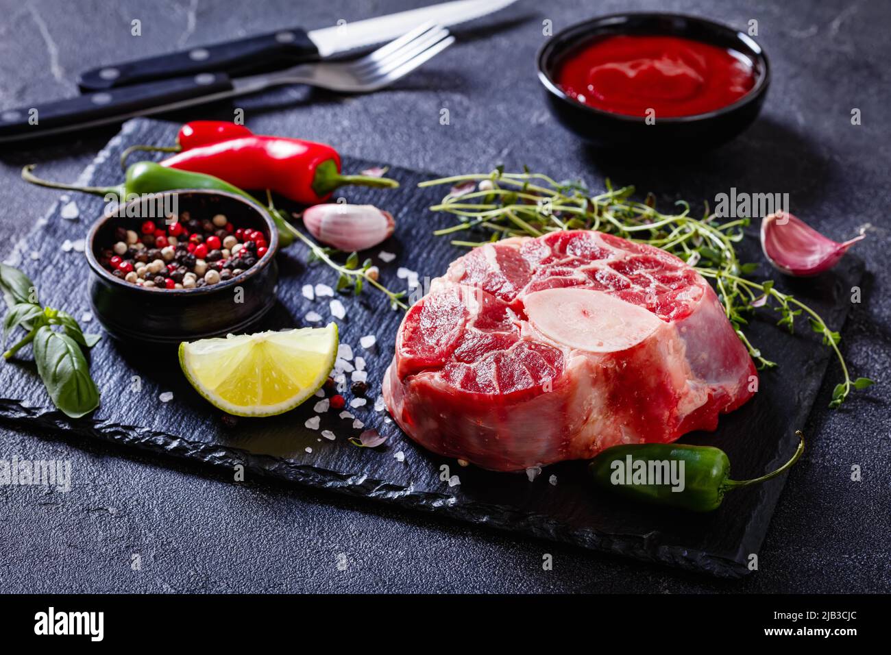 osso buco crudo, bistecca di vitello con marrowbone su tavola di pietra nera con timo, pepe in grani, timo fresco, peperoncino, aglio e lime su tavola di cemento Foto Stock