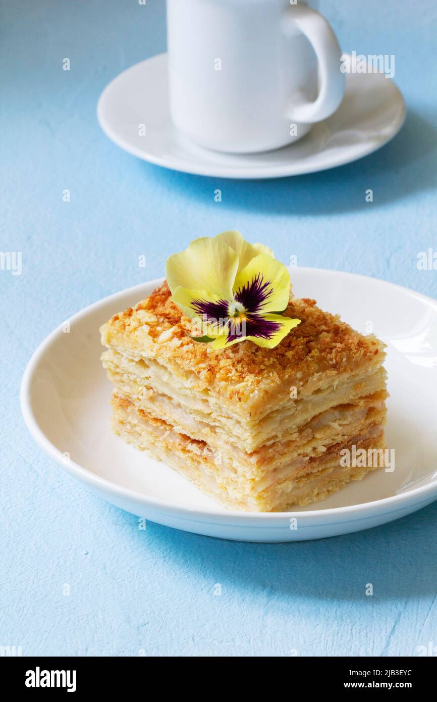 Una fetta di torta Napoleone, decorata con un fiore di viola, servita con una tazza di caffè o tè. Foto Stock