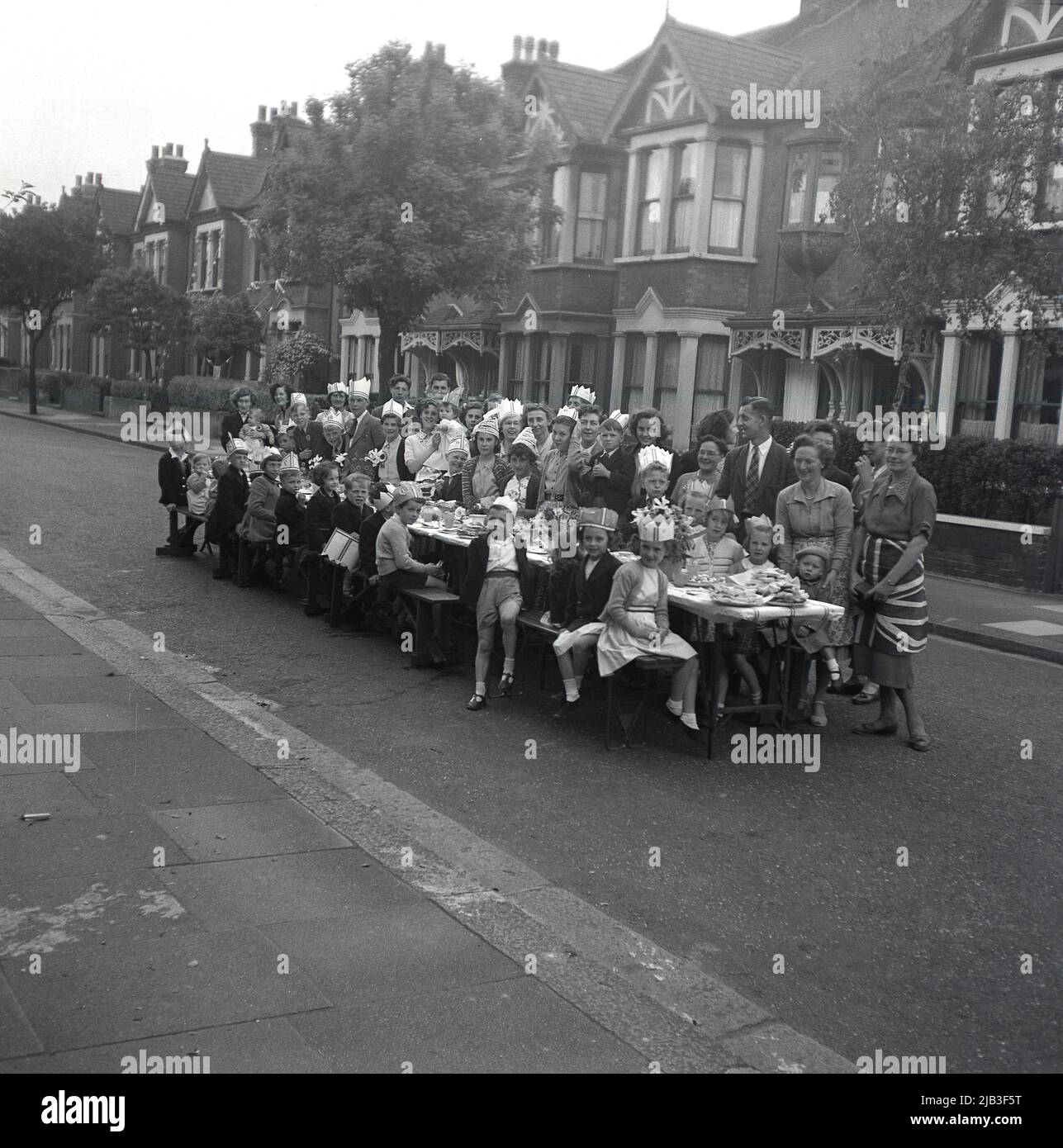 1953, storico, festa di strada, un gruppo di uomini, donne e bambini si radunano intorno ad un lungo tavolo per la strada per la loro foto, mentre celebrano l'incoronazione della regina Elisabetta II al trono, Inghilterra, Regno Unito. Come pochi nel dopoguerra avevano una televisione, persone e comunità sono scesi in strada per celebrare, un'occasione per le persone di tutte le età di riunirsi. Foto Stock