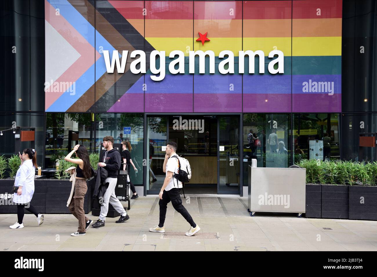 La gente cammina accanto al ristorante Wagamama in St Peter's Square, Manchester, Inghilterra, Regno Unito, Isole britanniche, Ora con la bandiera di avanzamento sul relativo fronte. La bandiera Progress Pride è stata sviluppata nel 2018 dall'artista Daniel Quasar. Basato sull'iconica bandiera arcobaleno del 1978, la riprogettazione celebra la diversità della comunità LGBTQ e richiede una società più inclusiva. Foto Stock