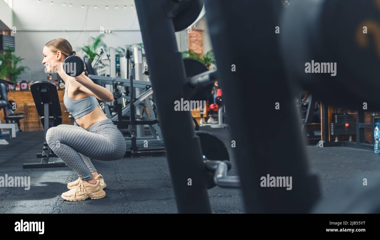 Brunette sottile persistente allegra donna nel suo 20s indossando abbigliamento sportivo grigio, sollevando un po 'di barbell leggero in una palestra moderna. . Foto di alta qualità Foto Stock