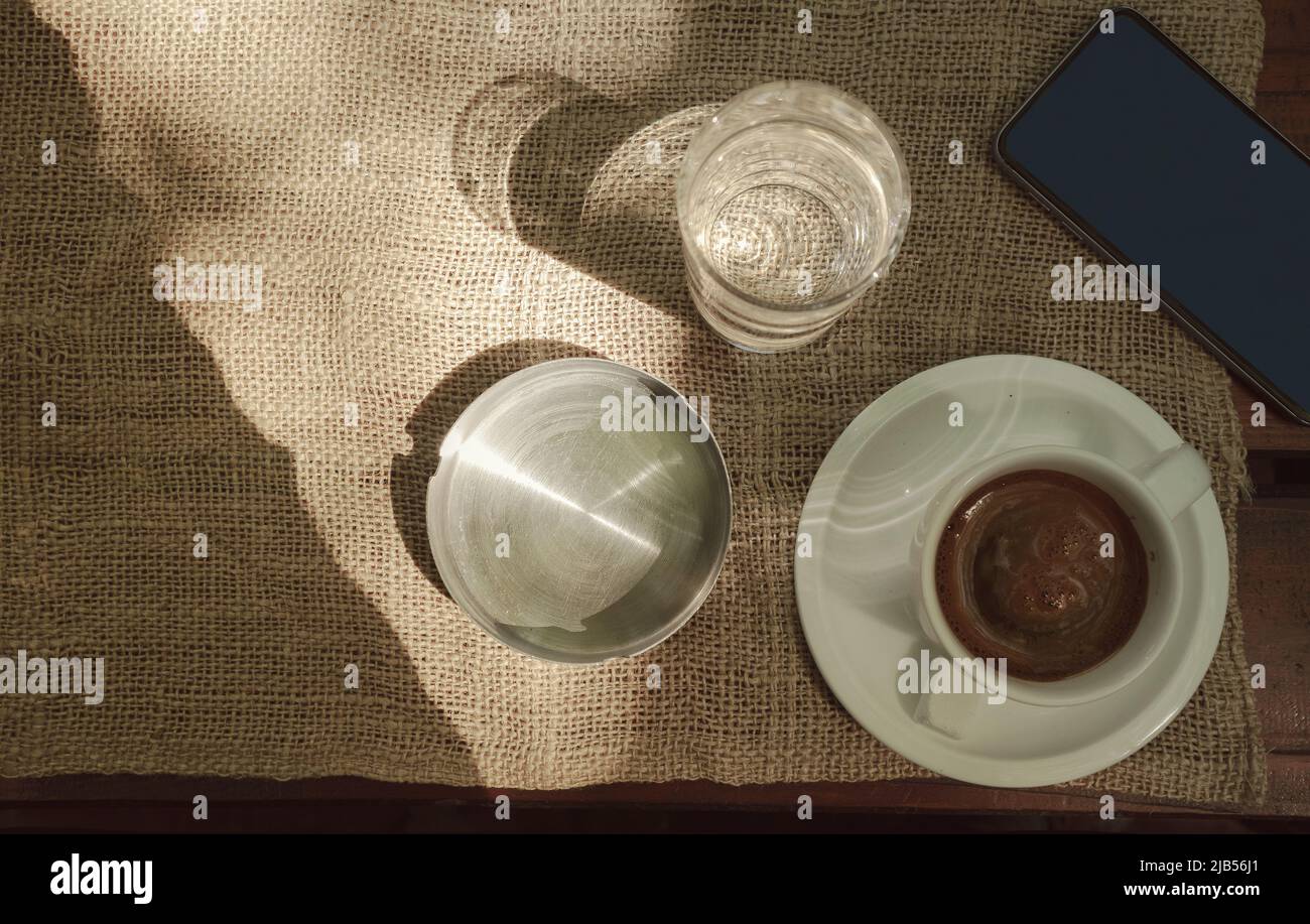 Tazza di caffè del mattino, bicchiere d'acqua, telefono cellulare e vassoio di cenere sul vecchio tavolo di legno, guardato dall'alto. Foto Stock