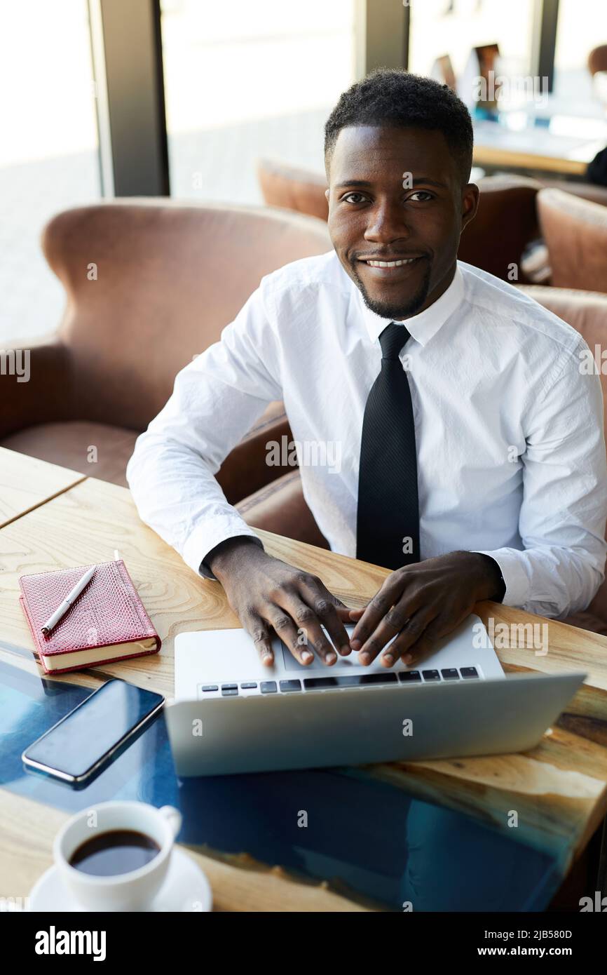 Contenuto sicuro bel giovane uomo d'affari afroamericano in camicia formale seduta al tavolo in caffetteria e di lavoro con file online su laptop Foto Stock
