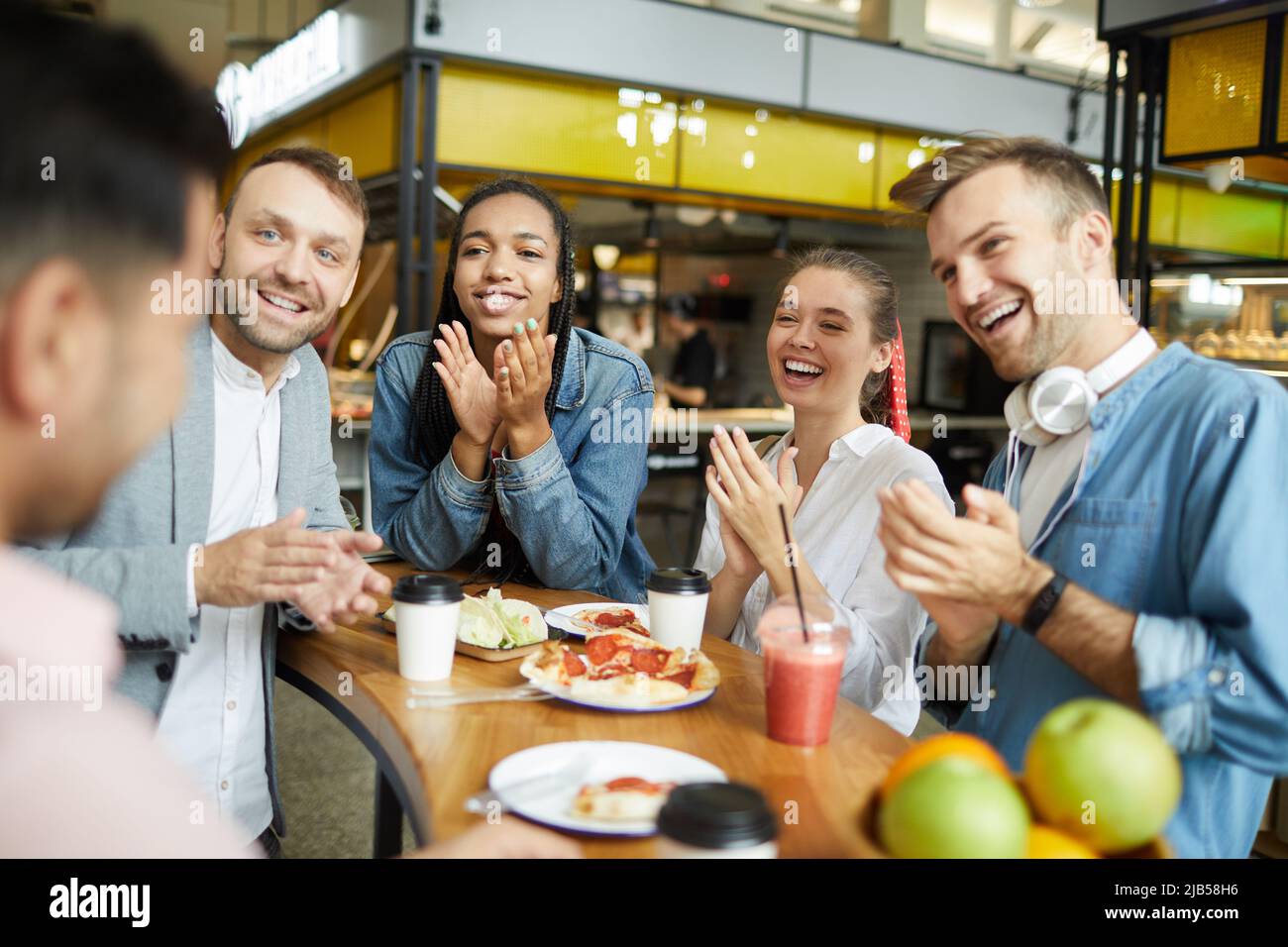 Gruppo di giovani positivi multietnici in abiti casual seduti a tavola con spuntini e applauding mentre si congratulano con l'amico in caffetteria Foto Stock