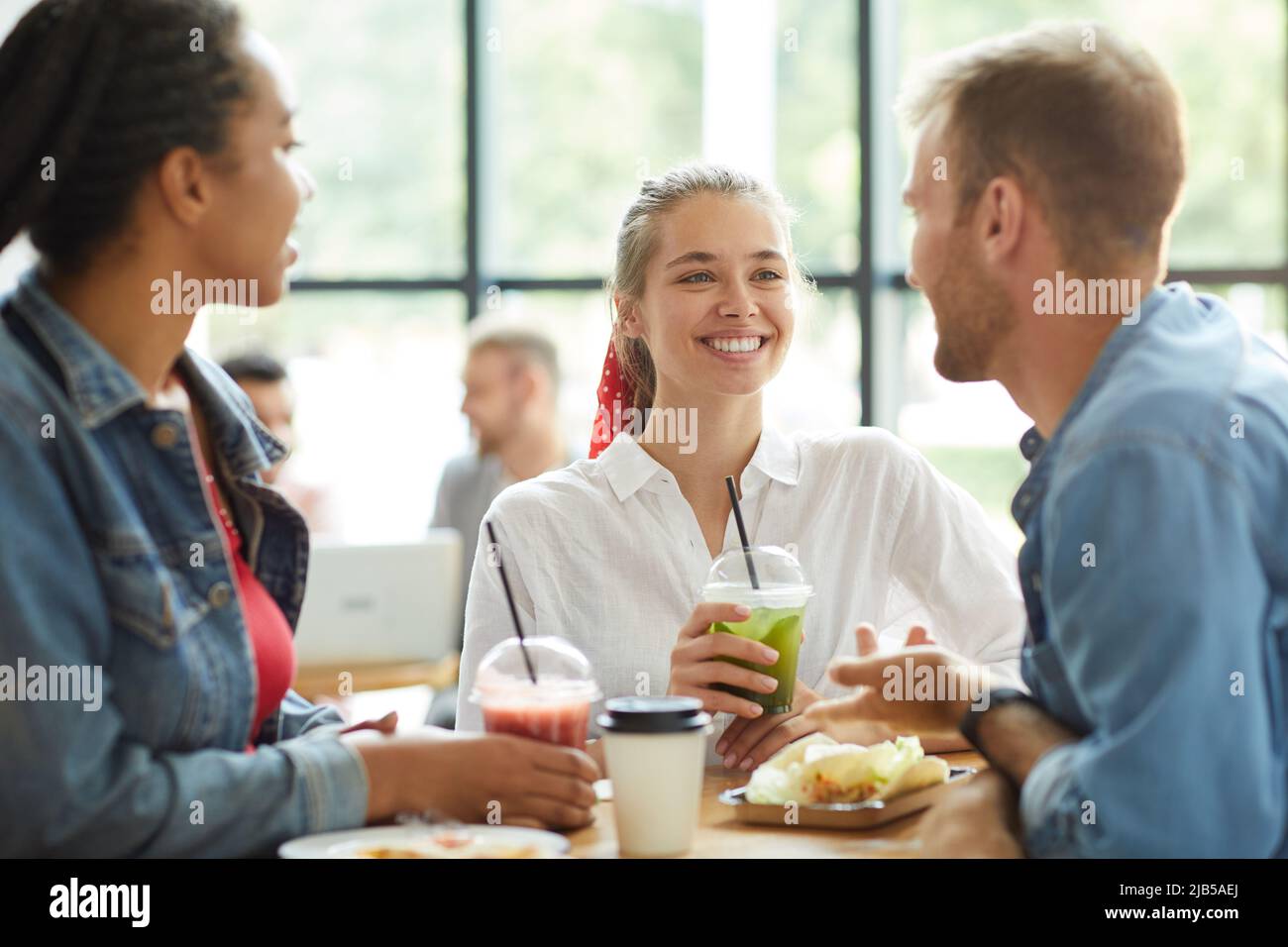 Gruppo di giovani amici multietnici positivi e spensierati in abiti casual in piedi al tavolo e pranzare insieme in un fast food cafe Foto Stock