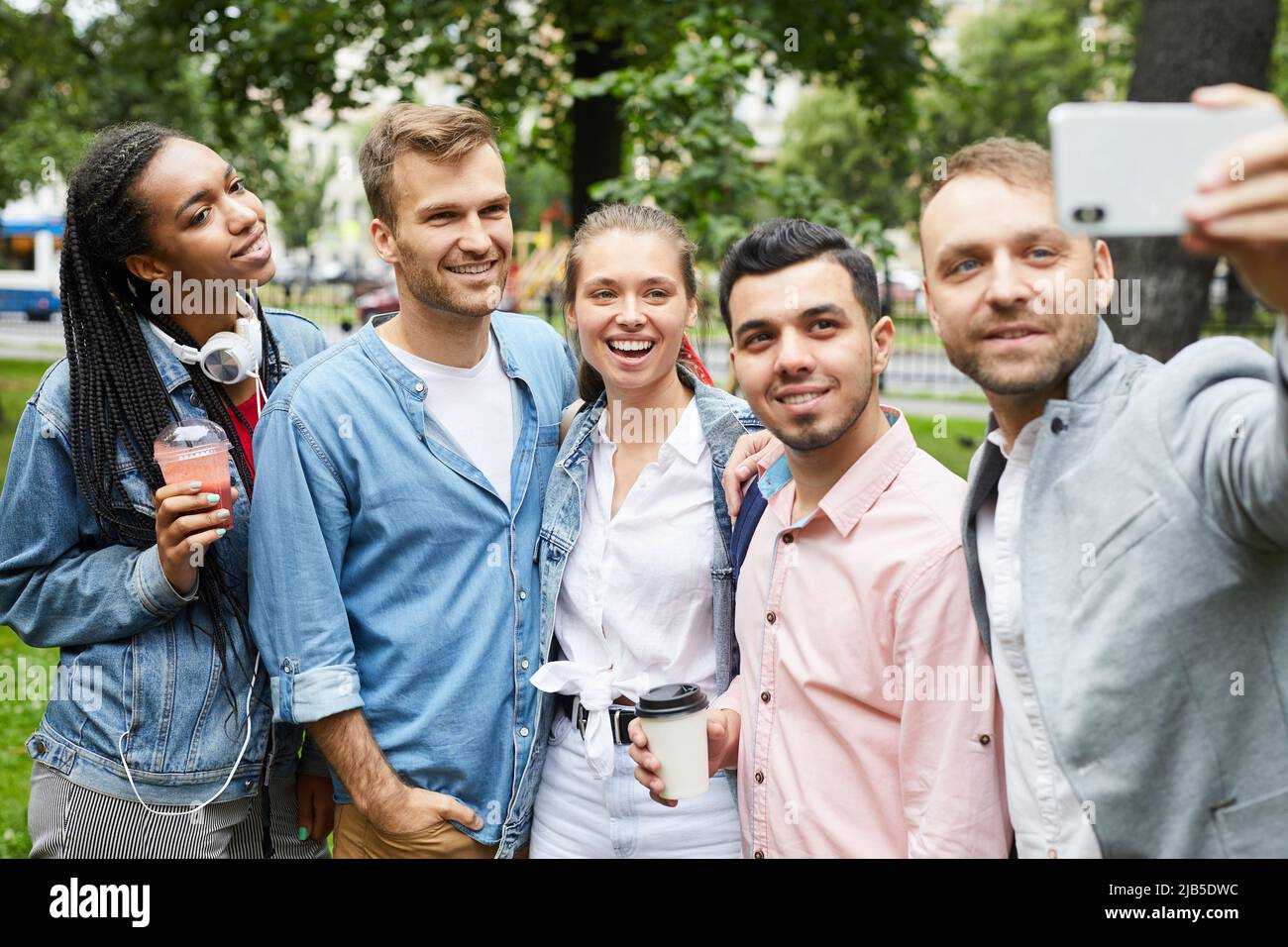 Gruppo di giovani positivi multietnici in abiti casual in piedi nel parco e fare autoritratto su smartphone Foto Stock