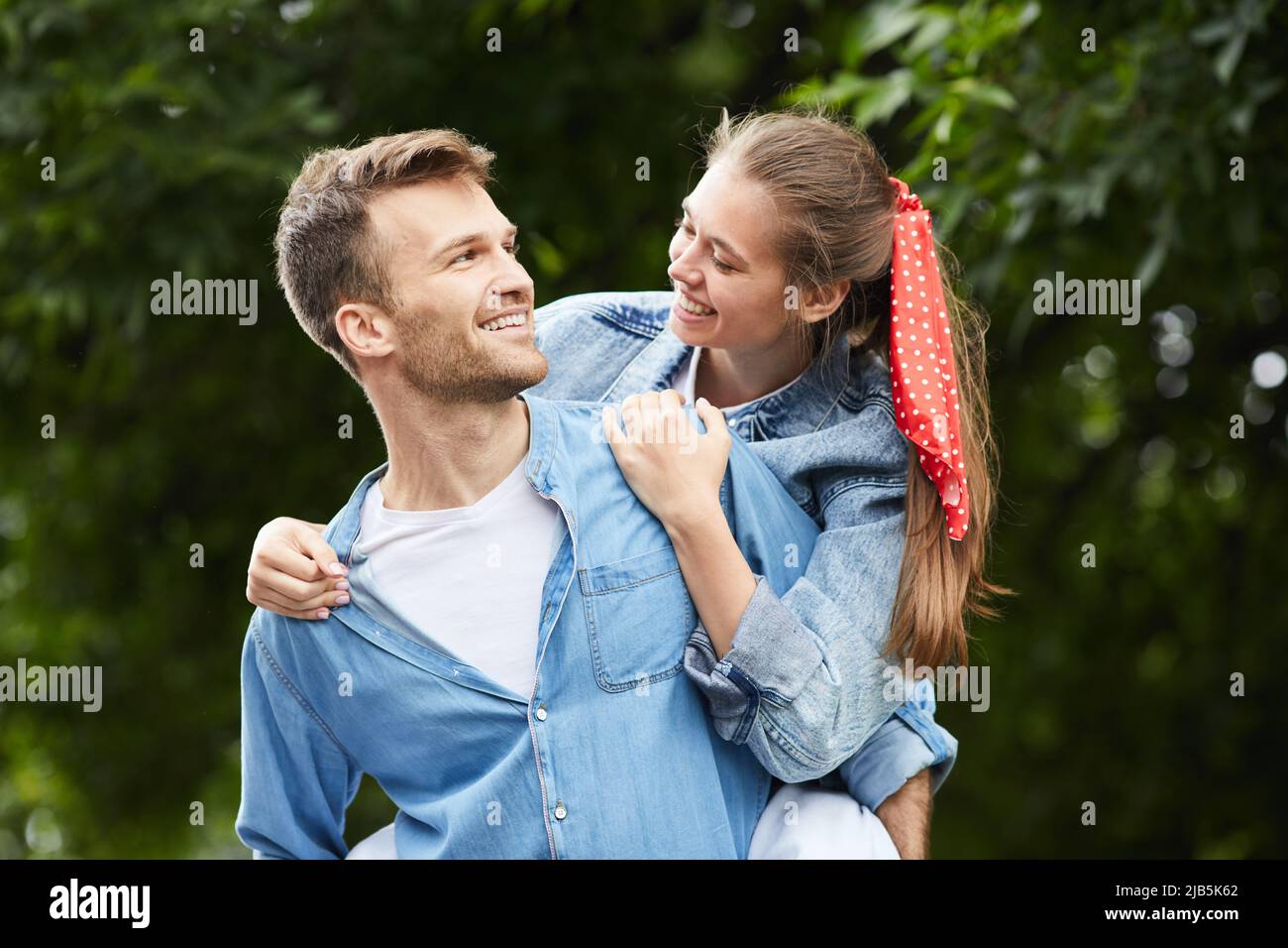 Positivo bel ragazzo giovane in denim camicia guardando la ragazza mentre dando piggyback a lei nel parco estivo Foto Stock