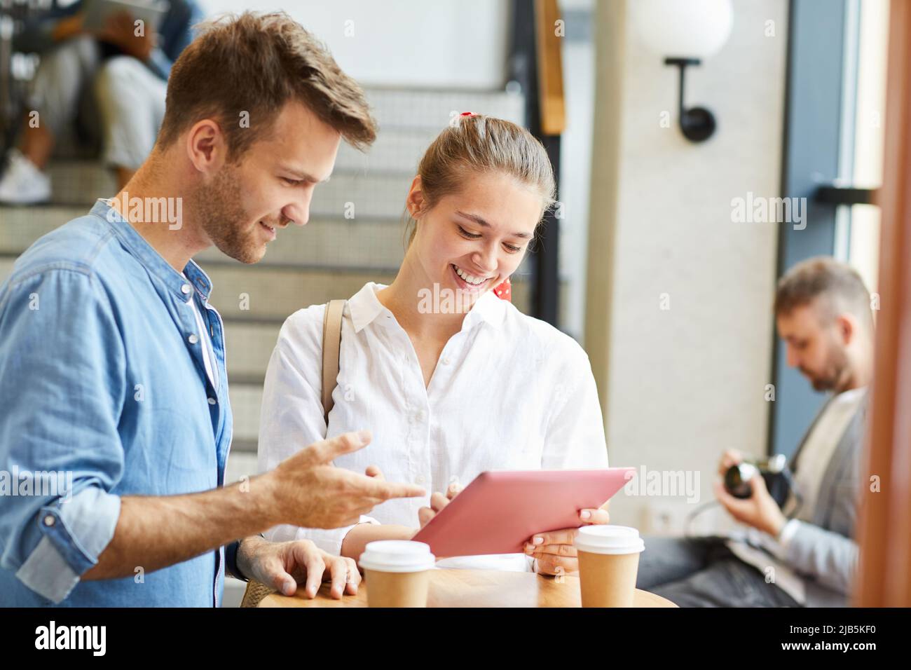 Giovani amici positivi in piedi nella caffetteria dell'università e usando il tablet digitale mentre discutono la piattaforma in linea Foto Stock