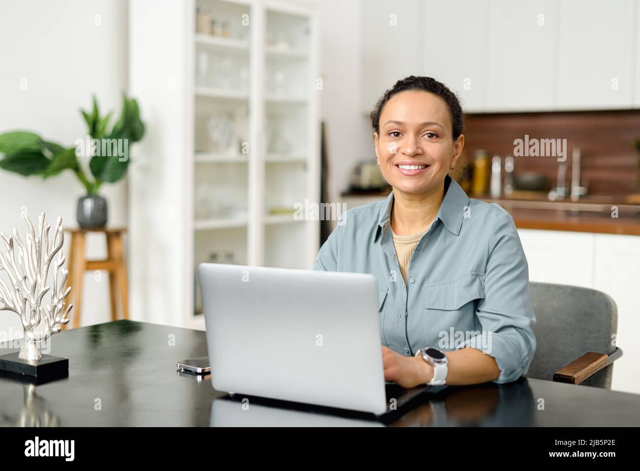 Dipendente femminile in abbigliamento casual elegante guarda la fotocamera e sorride, digitando messaggi, rispondendo alle e-mail, vista frontale. Donna di mezza età sicura lavora con un notebook presso l'ufficio moderno Foto Stock